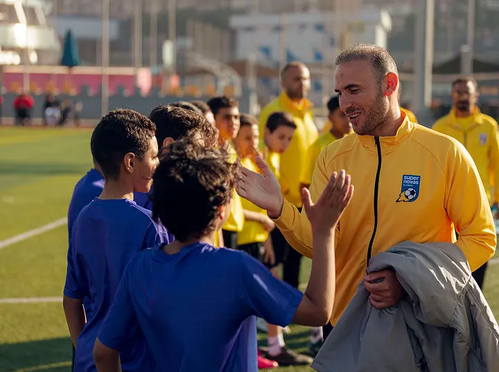 Coach in yellow jacket giving high-fives to kids in blue sports uniforms on a soccer field.