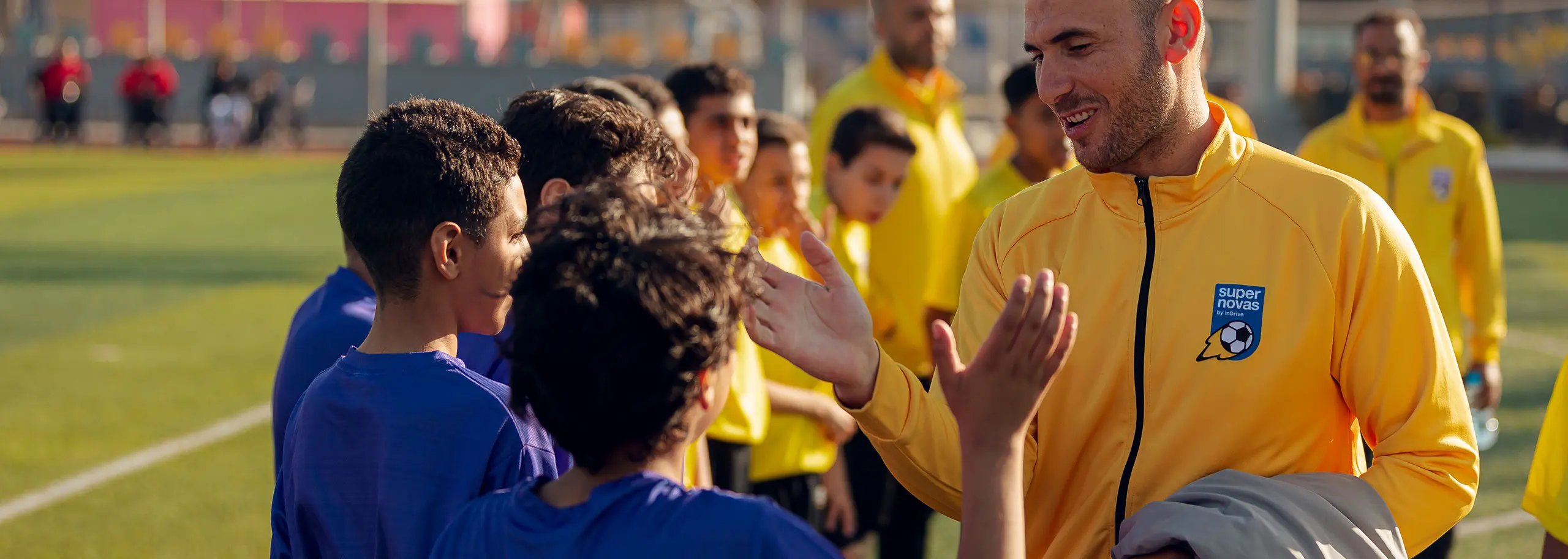 Coach in yellow jacket giving high fives to young soccer players in blue jerseys on a soccer field.
