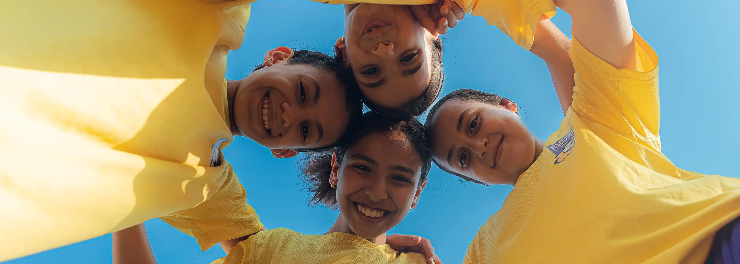 Four smiling girls wearing yellow shirts standing in a circle with heads close together against a blue sky.