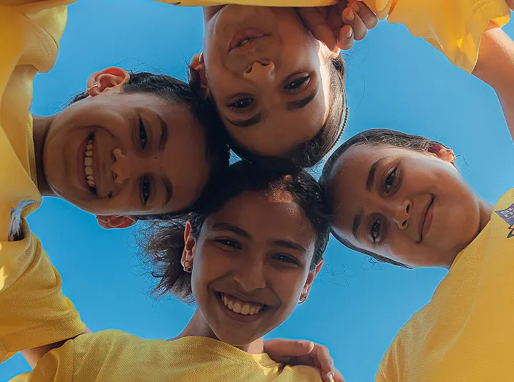 Four smiling young girls in yellow sports jerseys forming a circle and looking down at the camera against a clear blue sky.