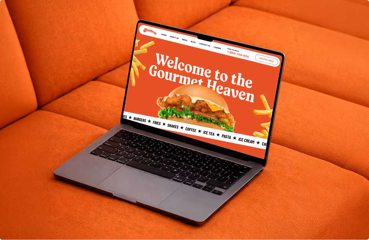 Laptop on orange couch displaying a website with a chicken sandwich and text 'Welcome to the Gourmet Heaven'.