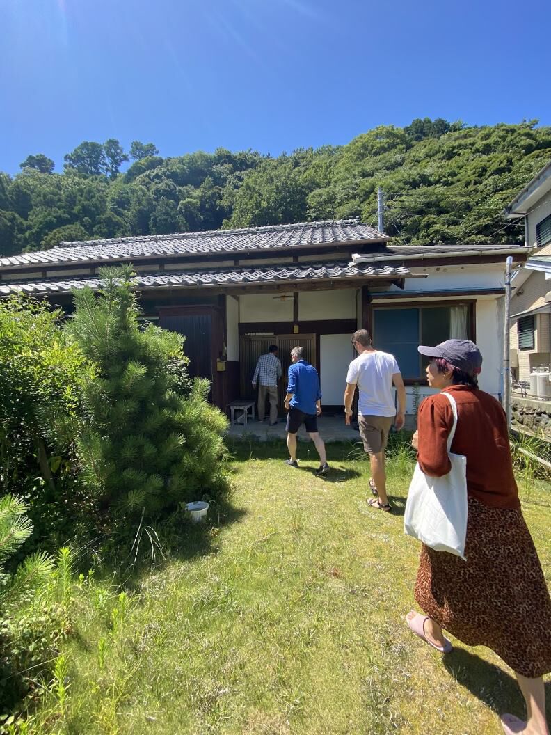 Visiting a Japanese home in the countryside. A group of 4 individuals is seen walking up toward a house that is nestled in the forest. There is a tree in the foreground and grass on the ground.