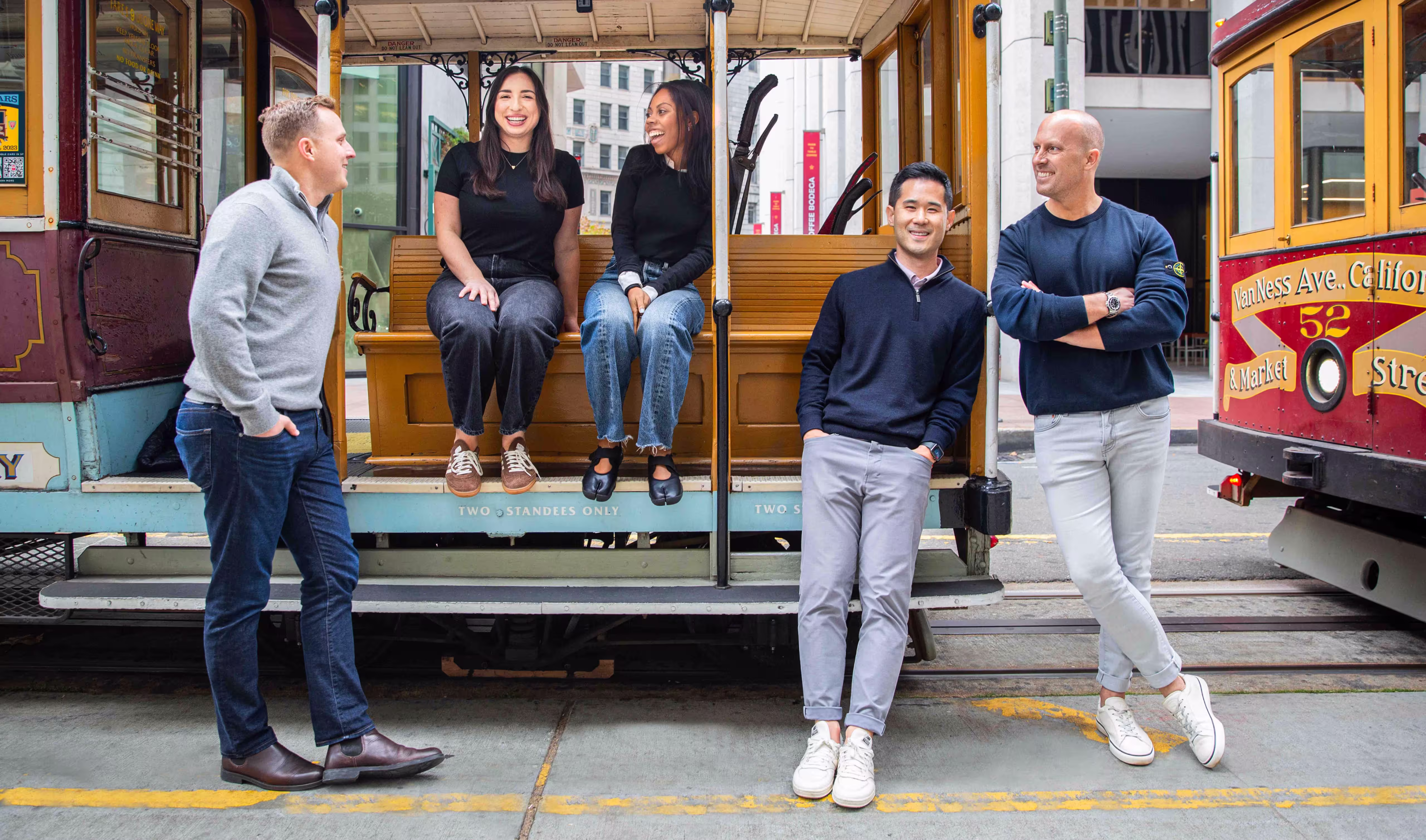 Five casually dressed people smiling and interacting while leaning against and sitting on a vintage cable car in an urban setting.