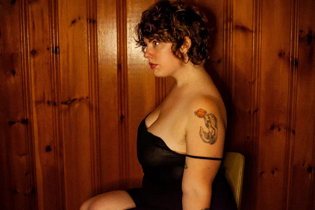 Profile of Jill, sitting up straight with her eyes closed, wearing a black tank top with a strap falling down, against a wood paneled wall.