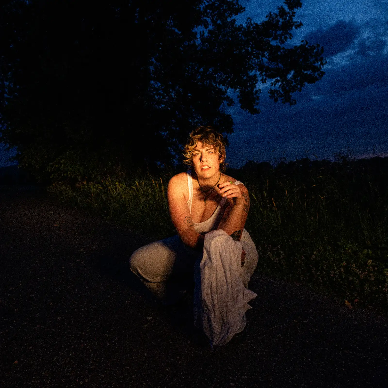 A wide night portrait of Jill in a light outfit kneeling in front a tree with a golden light shining into their face