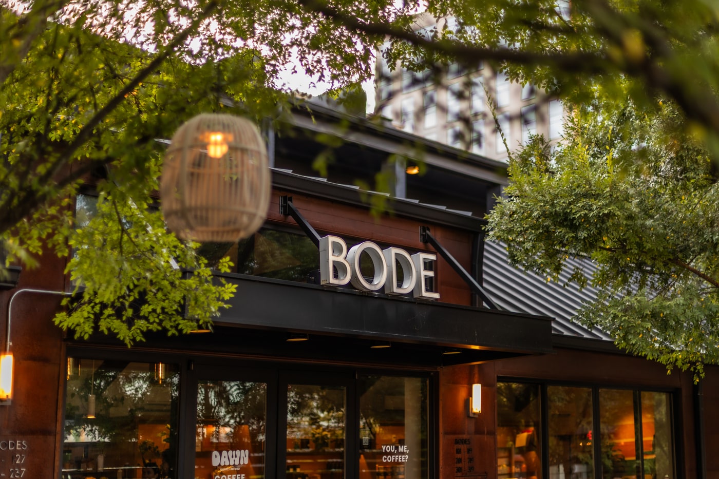 BODE hotel entrance with DAWN coffee shop sign under leafy trees, warm lights glowing on the street.