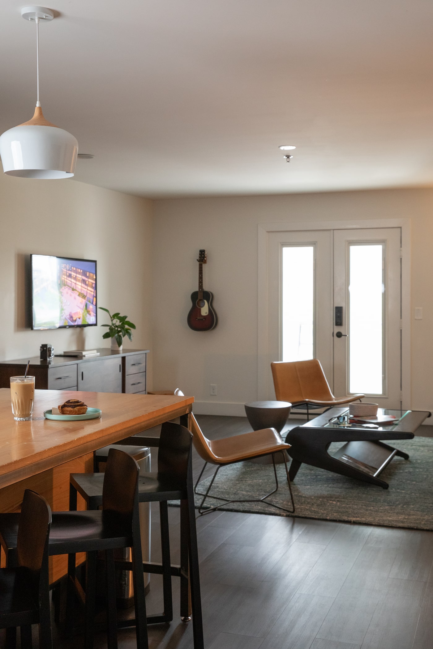 Modern open-plan hotel suite with kitchen island, lounge chairs, wall-mounted TV, and bright patio doors.