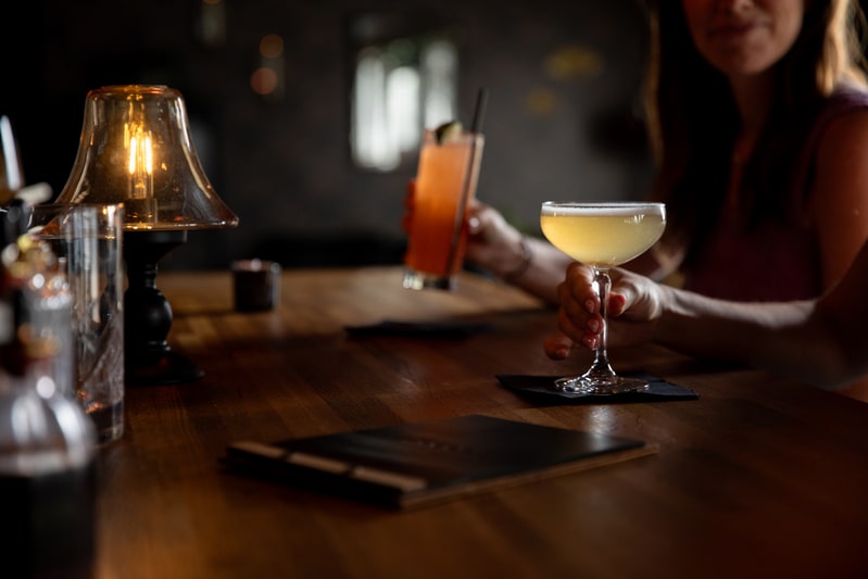 Candlelit bar scene with cocktails at BODE, guest holding drinks over a wooden counter.