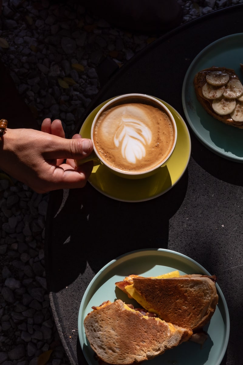 Latte with leaf art on sunny patio table with breakfast sandwich, toast and banana tart.