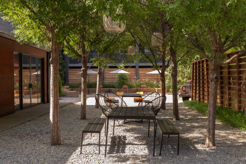 Shaded courtyard patio with metal table and benches under trees at BODE, outdoor seating area.