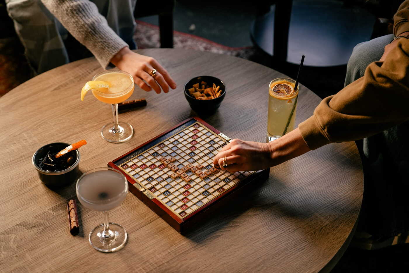 Two people playing Scrabble on a round wooden table surrounded by cocktails and snack bowls at a cozy bar setting.