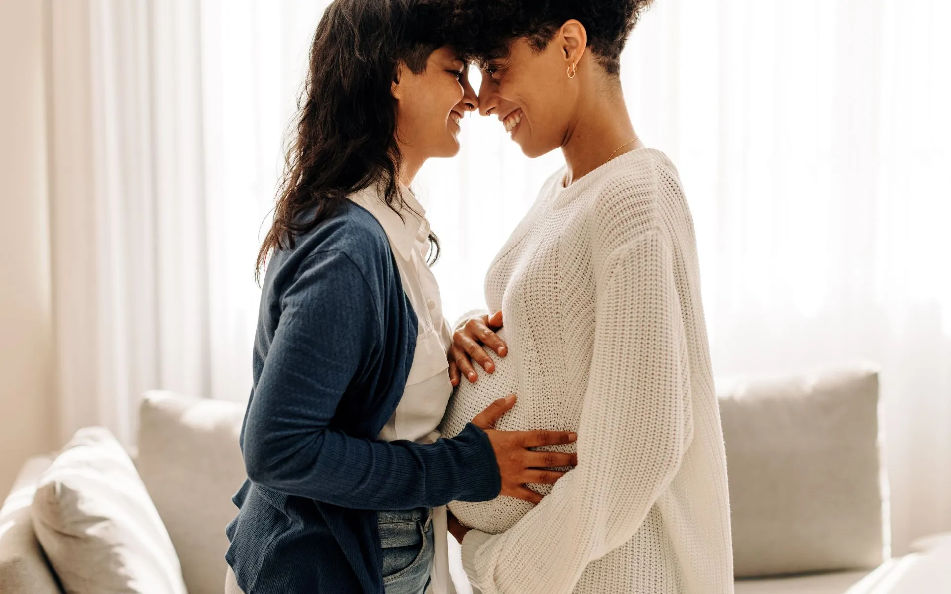 Two mothers stand close together, smiling at each other. The non-birthing partner holds the bump of the pregnant partner.