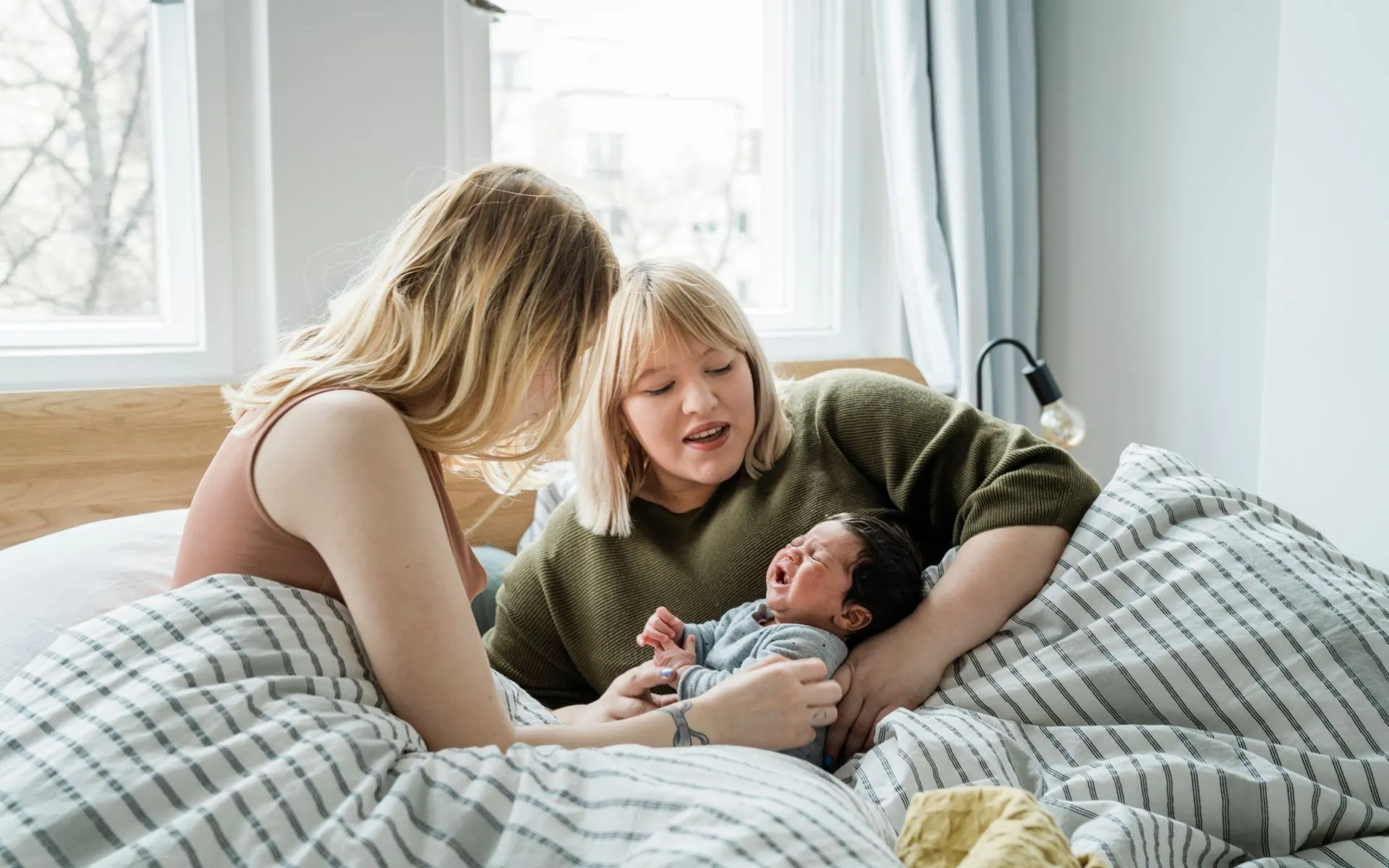 Two mothers sit in bed speaking and cradling their baby. 