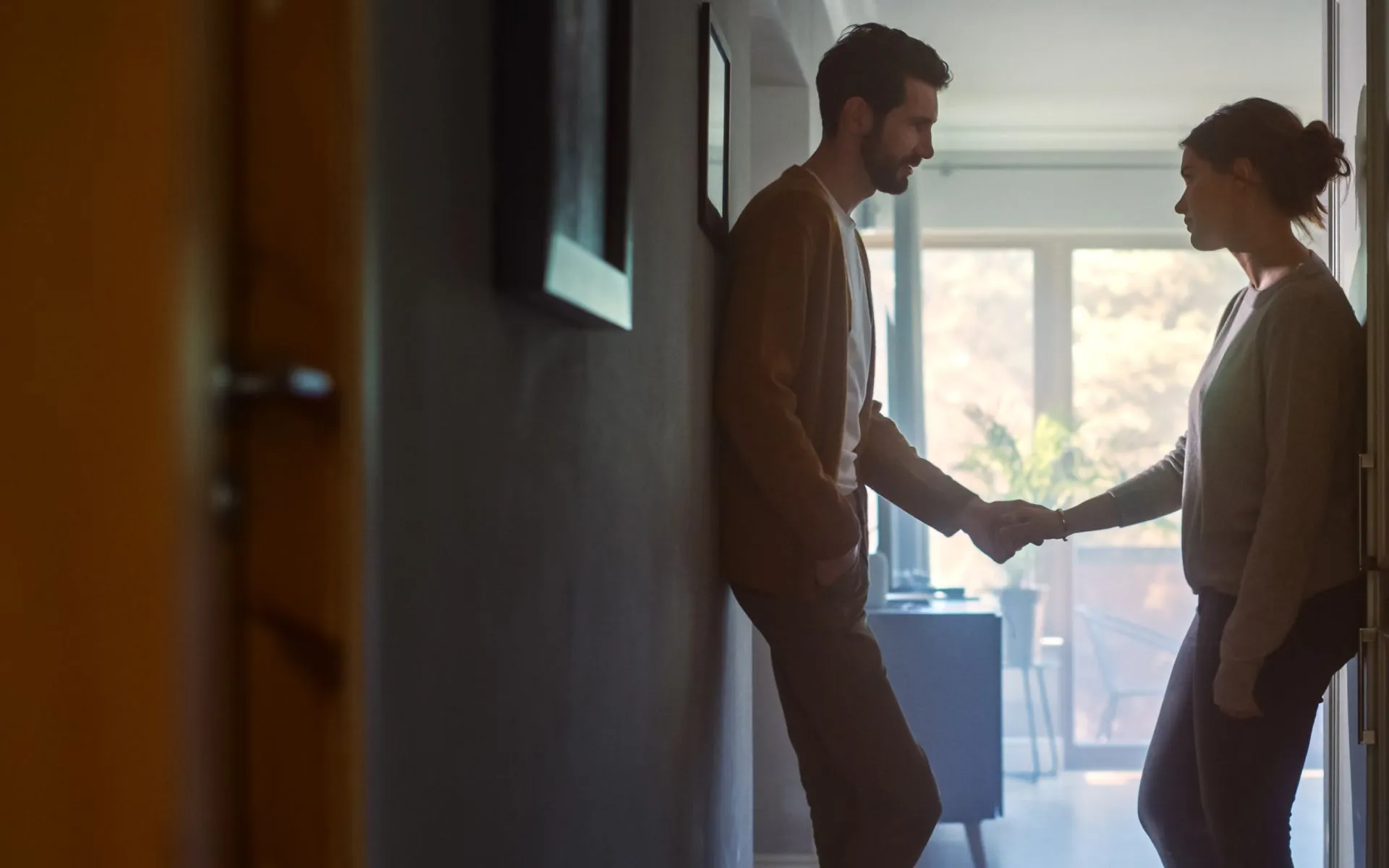 A couple stand in the hallway of their home. They each other's hands while having a serious conversation.