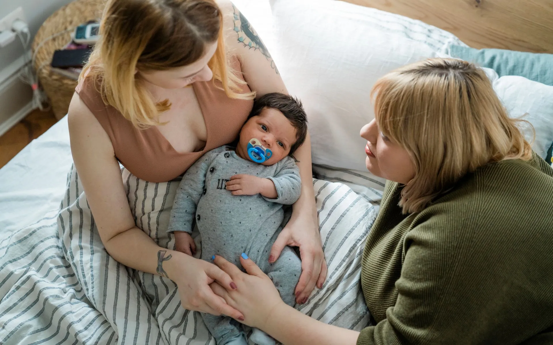 A mother sits cross-legged in bed with her baby in her lap. Her partner lays next to her, holding her hand. 