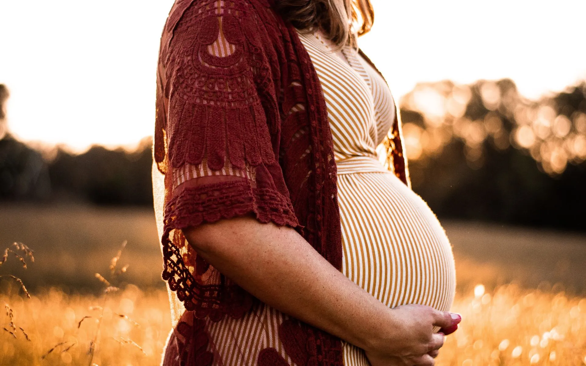 Heavily pregnant mother stands outside at sunset holding her bump.