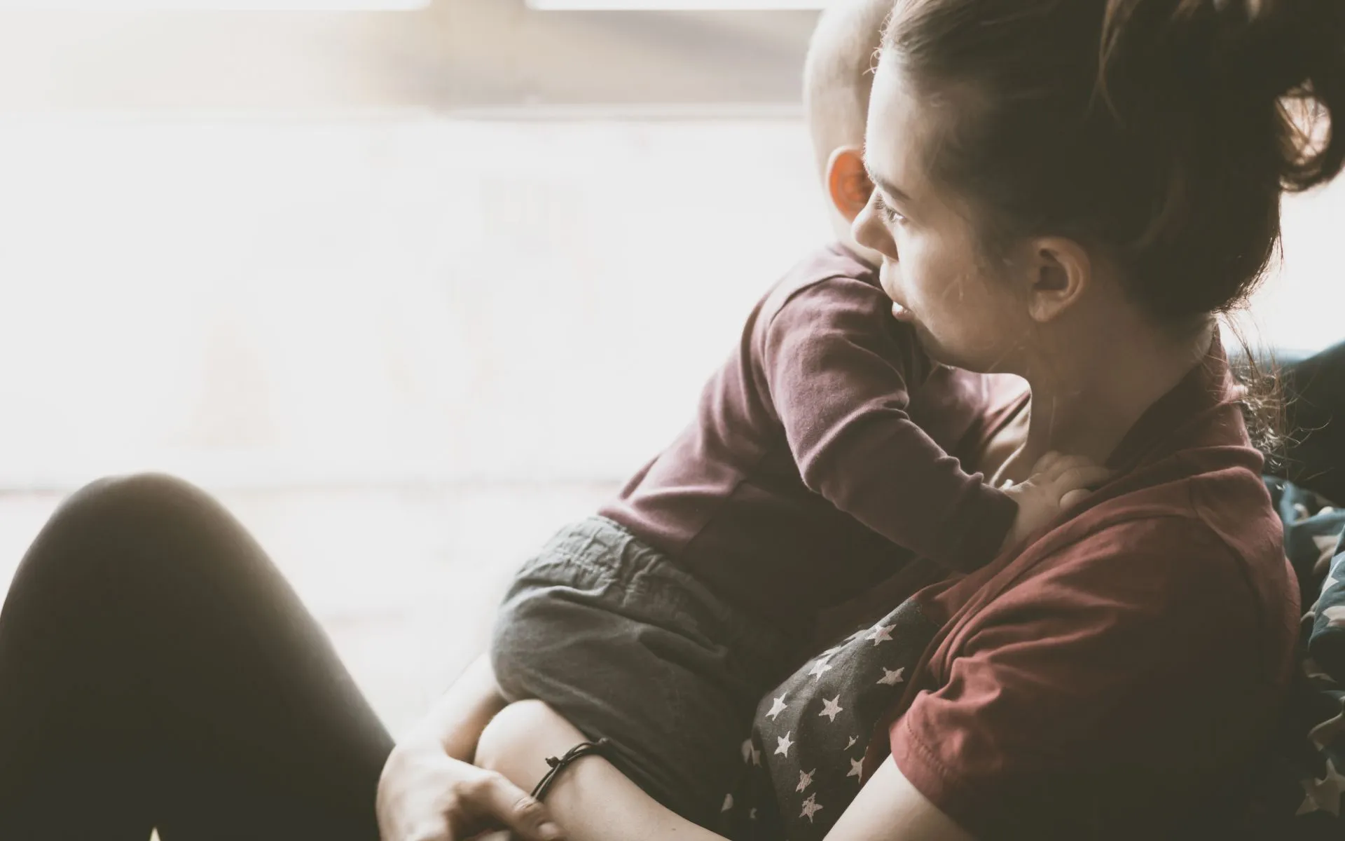 A mother sits on the floor with her back to the wall. Her young baby sits in her lap. 