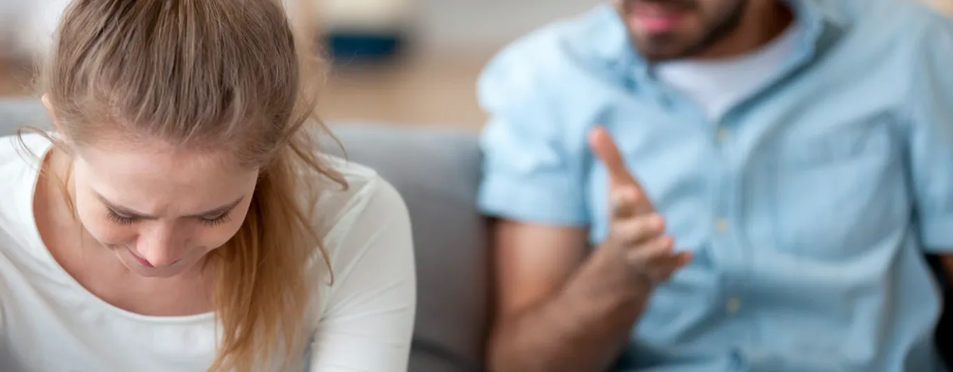 A man and woman on a couch, the man looking at the woman angrily, capturing a moment that suggests underlying coercive control.