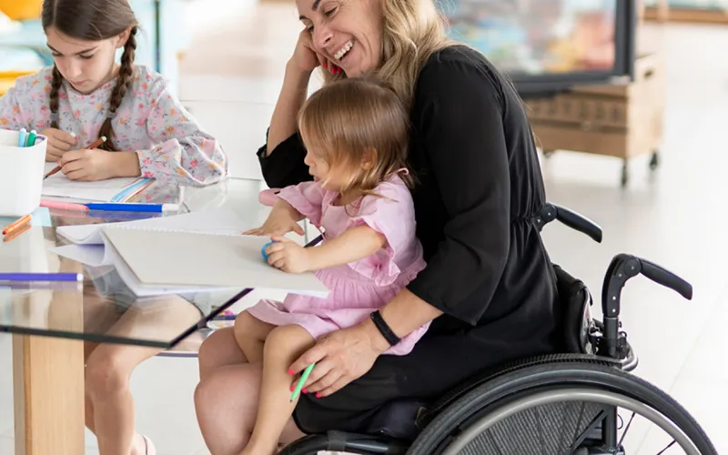 A mother sits in a wheelchair with her youngest daughter in her lap and another daughter sitting at the table with them. They smile while doing an art activity together. 