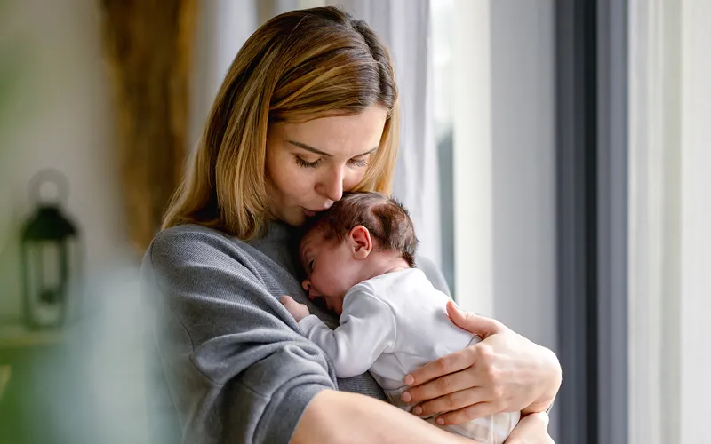 A mother hold her newborn to her chest and kisses the baby's forehead.
