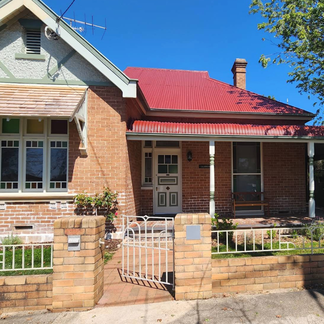 A charming brick house featuring a red roof, enclosed by a white fence, set against a clear blue sky.