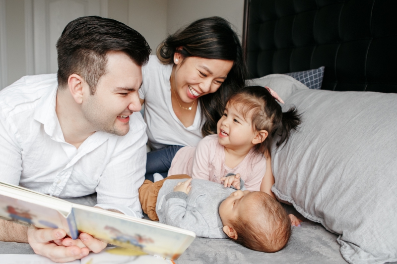 A diverse family of four, including a man, woman, baby, and toddler, happily reading a book together.