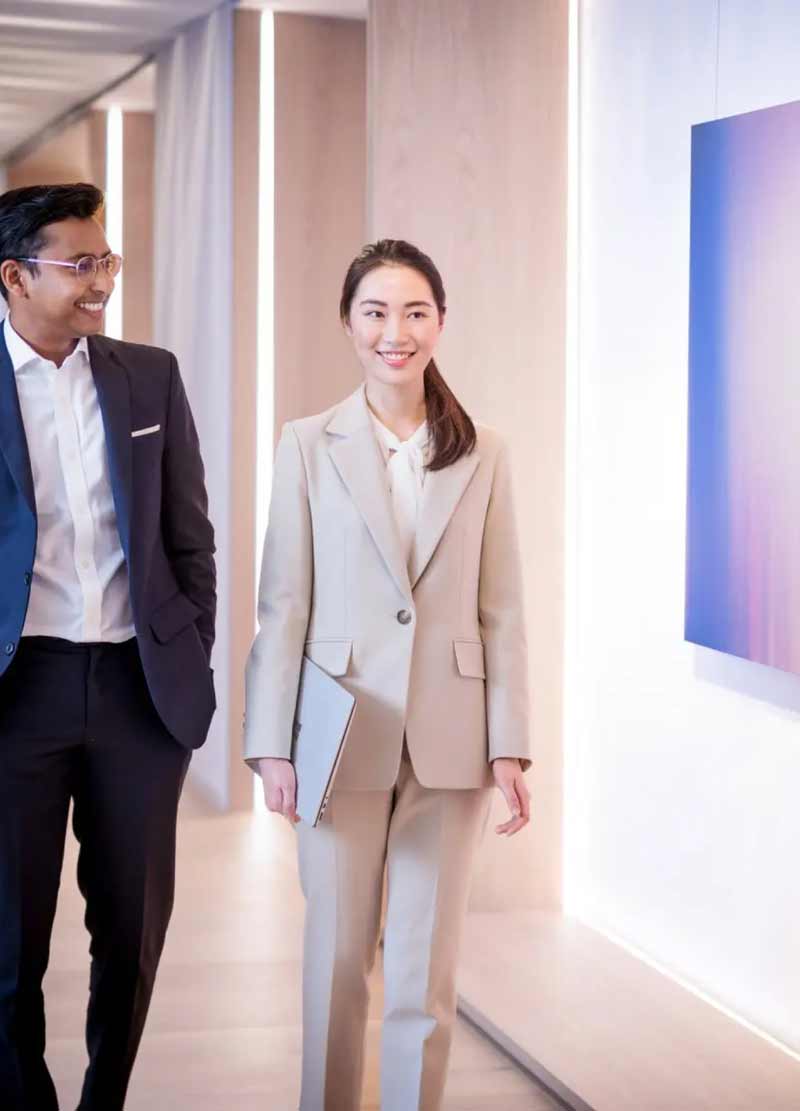Two professionals in suits walking indoors, woman holding a laptop and smiling ahead.