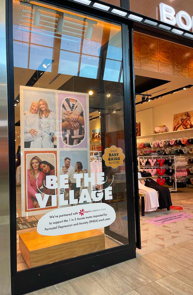 Storefront window with a 'Be The Village' sign supporting Aussie mums with perinatal depression and anxiety.