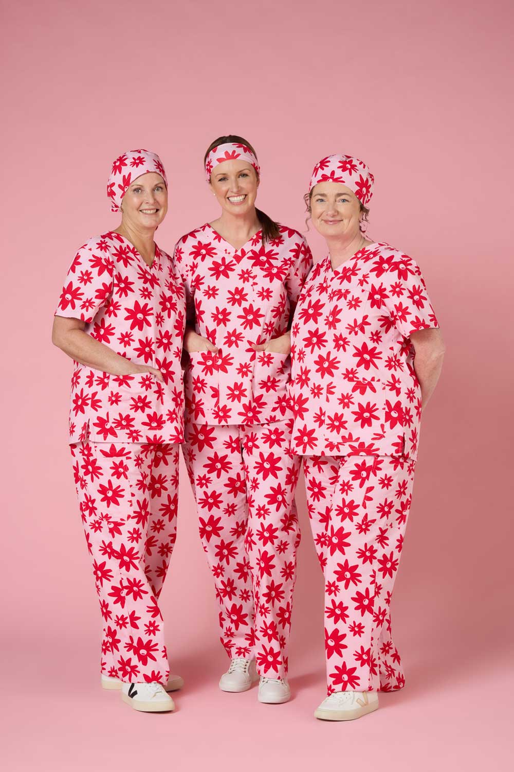Three smiling women wearing matching pink scrubs with red flower patterns and white shoes on pink background.