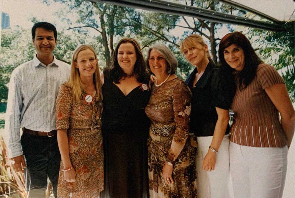 Group portrait of founders, ceo and family standing closely outdoors under a canopy with trees behind them.
