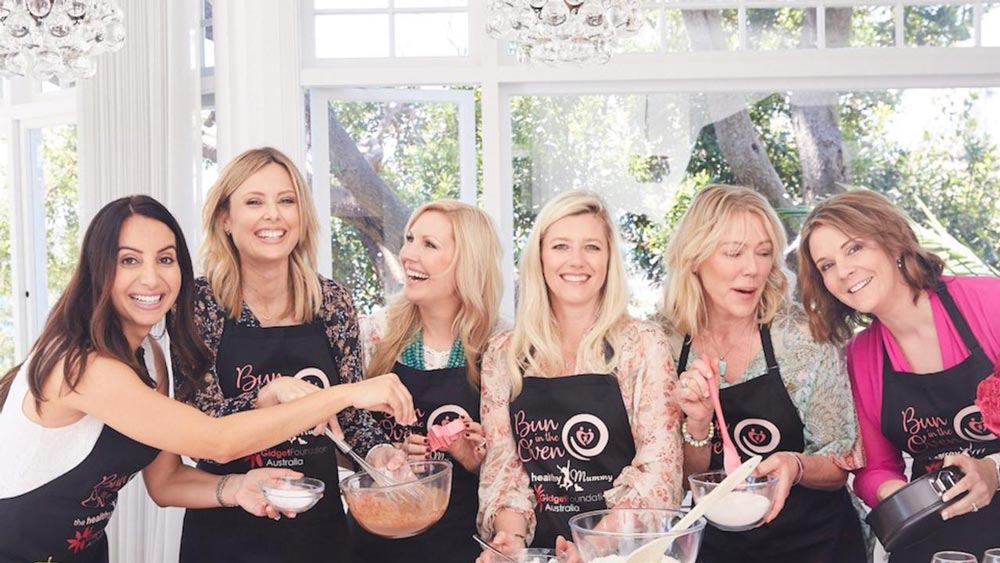 Six smiling women wearing aprons preparing baking ingredients in a bright kitchen.