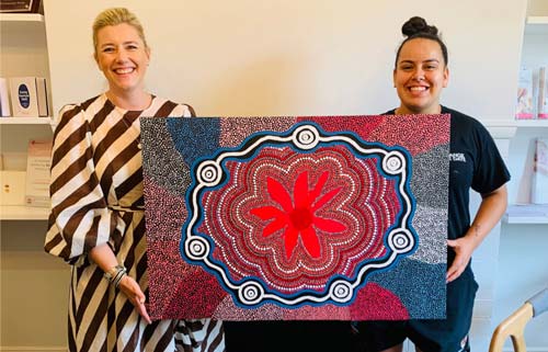 Two smiling women holding a colorful Aboriginal dot painting with red, blue, and white patterns.