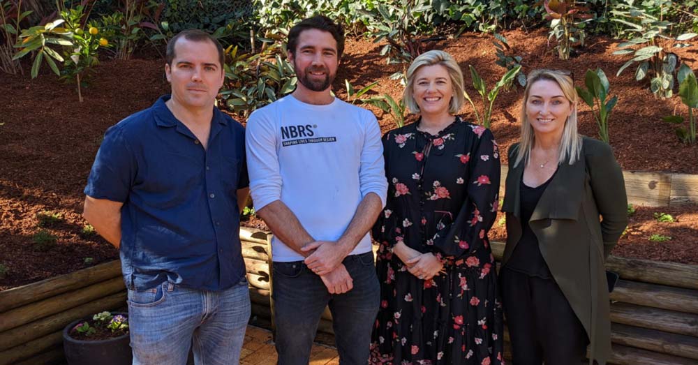 Four people standing outdoors in front of a garden bed with plants and mulch, smiling at the camera.