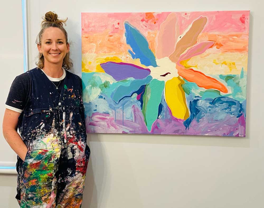 Smiling Meg Minkley in paint-splattered overalls stands next to colorful abstract flower painting on wall.