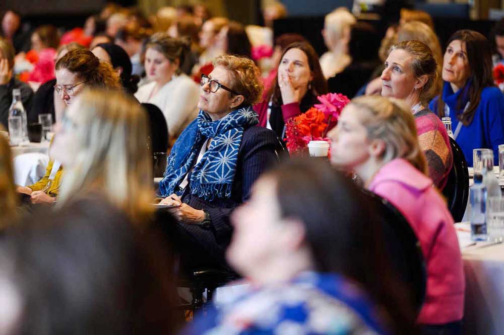 Audience attentively listening at a conference with colourful flowers on tables.