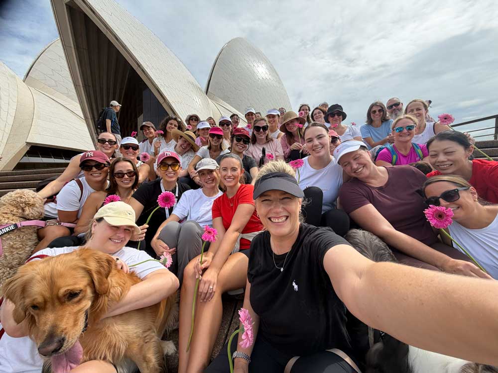 Team selfie sitting on steps with pink gerberas, and Sydney Opera House behind.