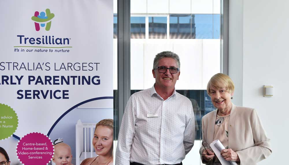 Two smiling older adults standing indoors near a window and a Tresillian early parenting service banner.
