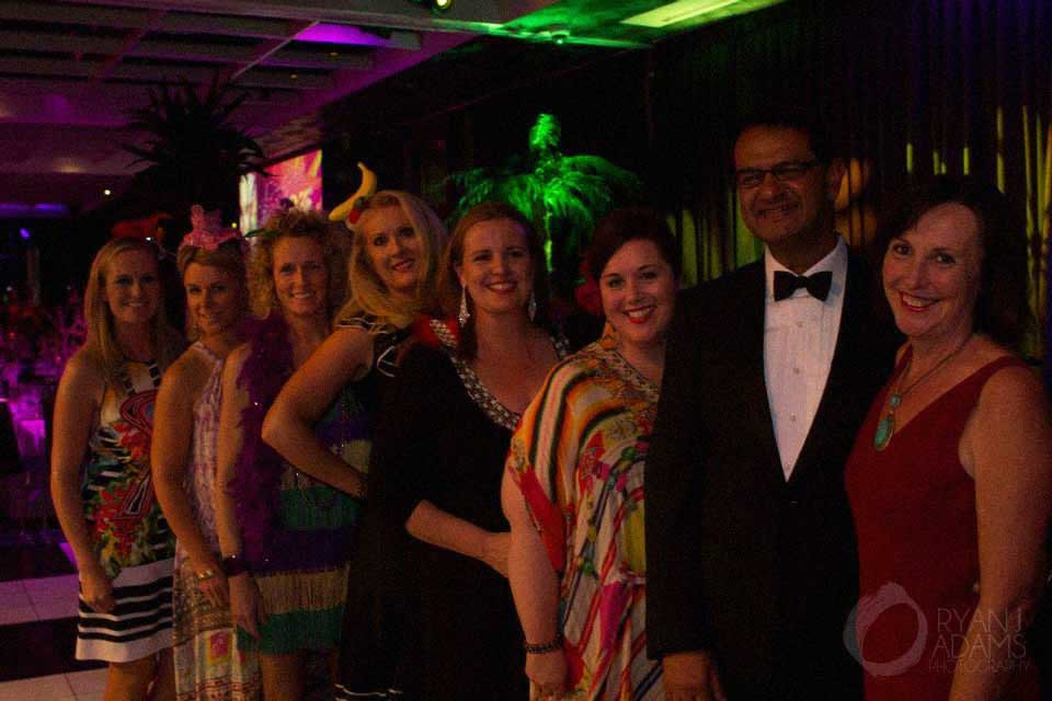 Group of seven people dressed in colorful dresses and a tuxedo smiling indoors at an event.
