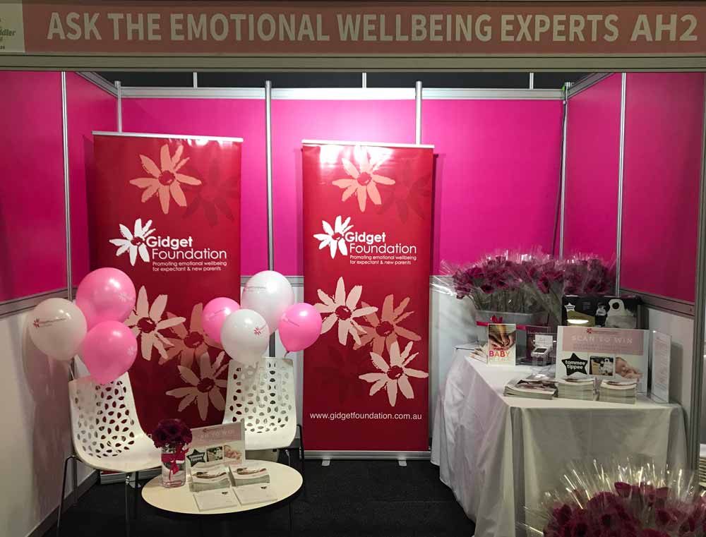 Booth with Gidget Foundation banners, pink and white balloons, brochures, and flower bouquets promoting emotional wellbeing.