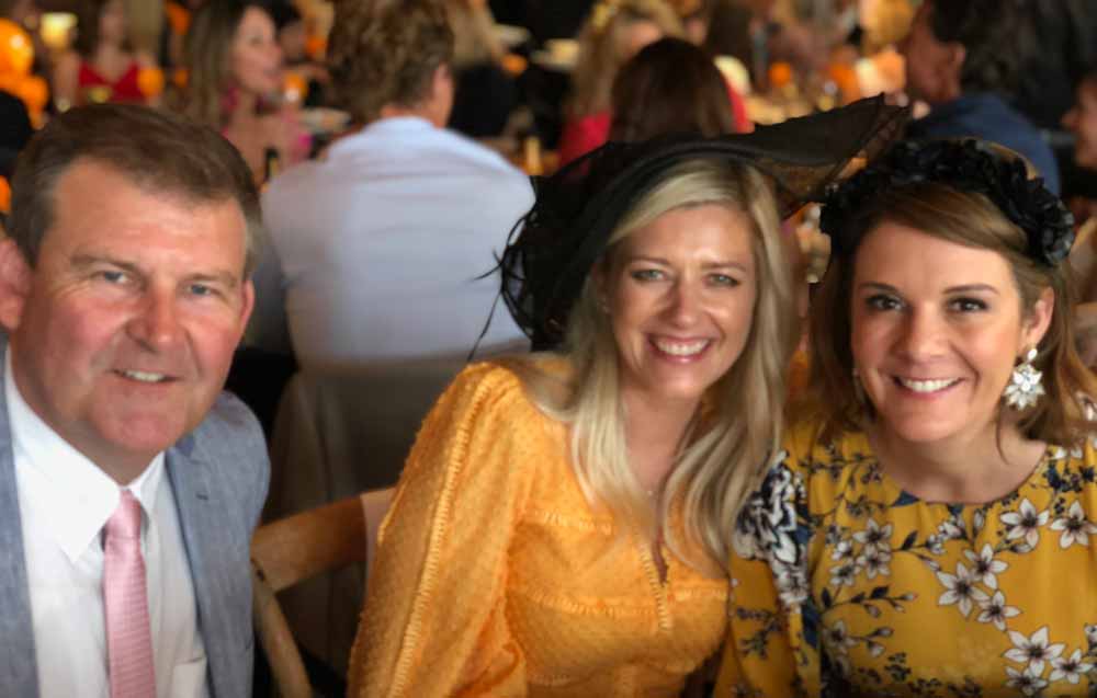 Three people smiling at a social event, with two women wearing floral dresses and headpieces.