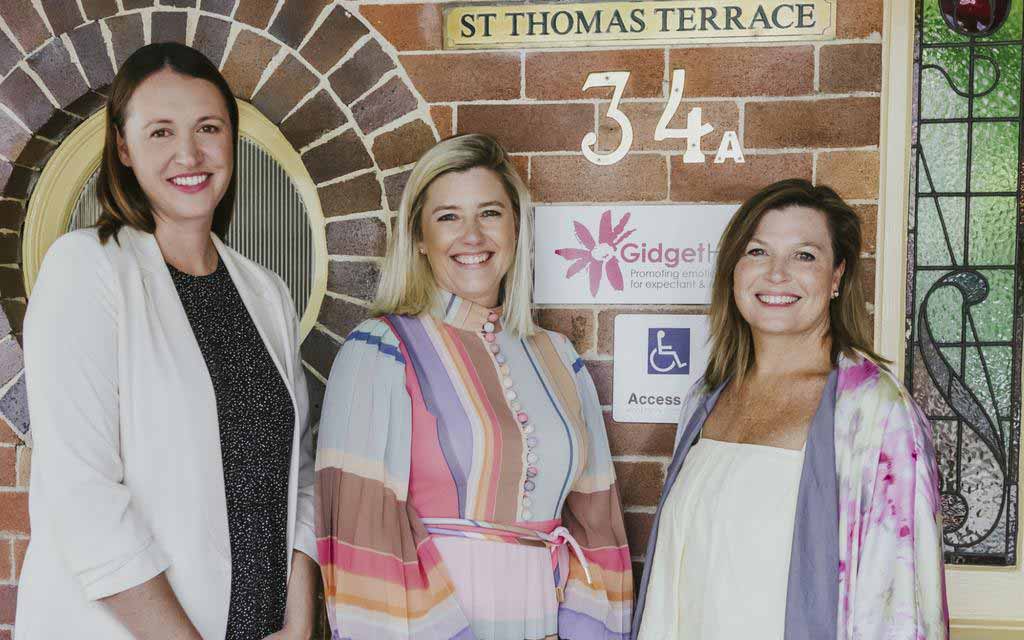 Three women standing and smiling in front of a brick wall with a St Thomas Terrace sign and Gidget House logo.