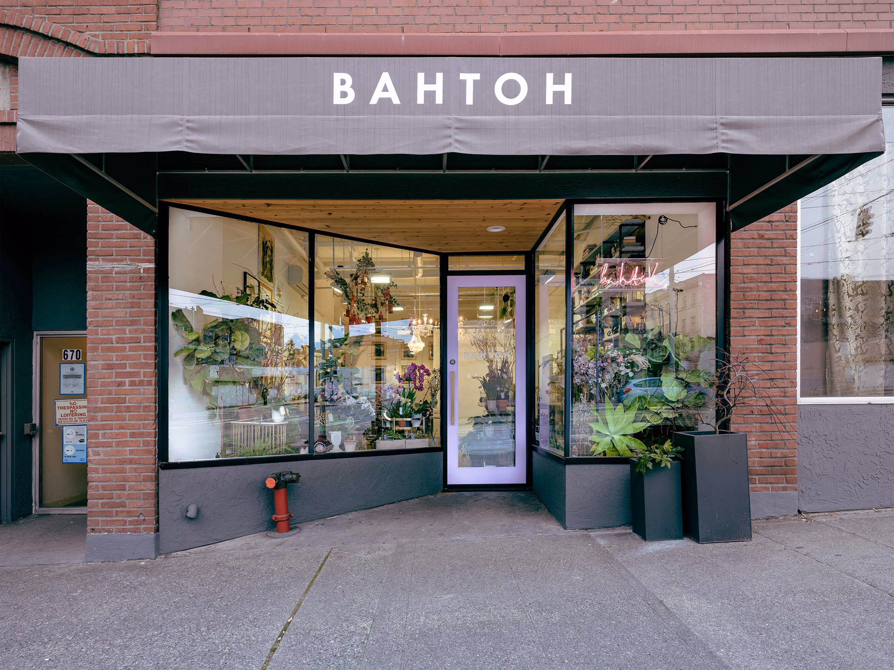 Exterior of BAHTOH salon, a brick storefront with a gray awning reading 'BAHTOH,' large display windows filled with plants and flowers, and a neon sign glowing inside