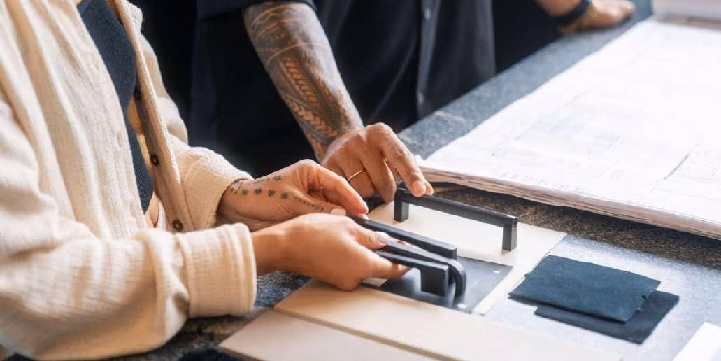 Close-up of two people's hands selecting cabinet hardware samples over architectural drawings on a table