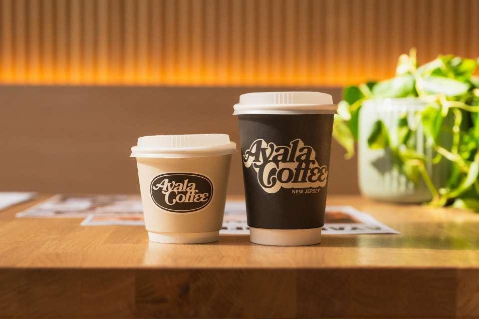 Two Ayala Coffee to-go cups with the shop's logo, sitting on a wood counter beside a plant