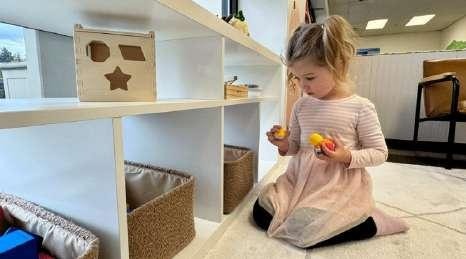 Child kneeling by open wooden shelving, playing with small toys at Laro Tayo