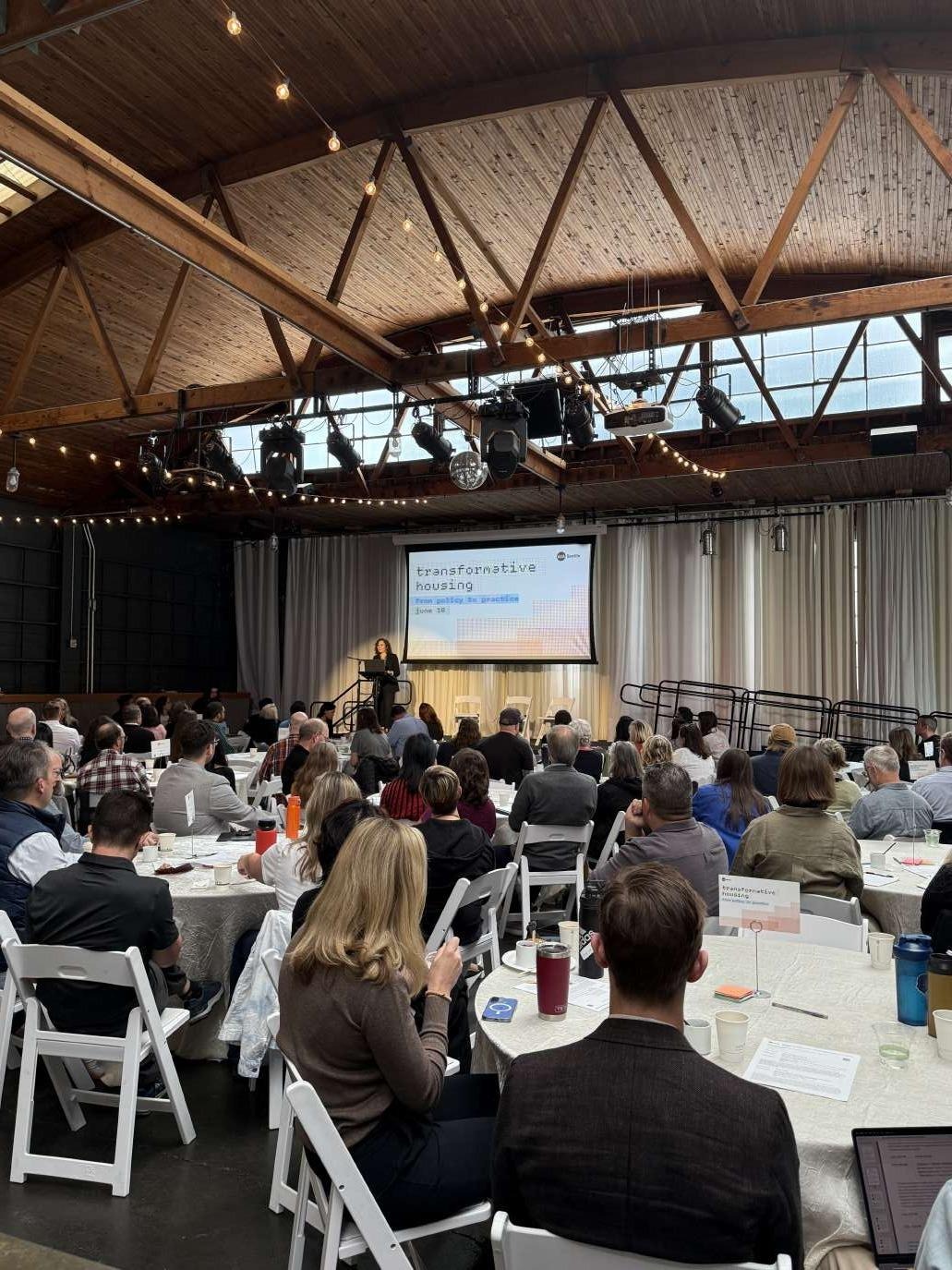 Audience seated at round tables listening to a speaker on stage under a wood-beamed ceiling with string lights, at a 'Transformative Housing' forum