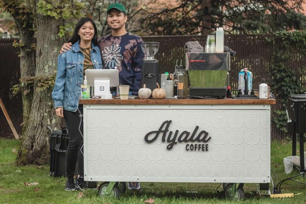 Two people smiling behind a mobile Ayala Coffee cart set up outdoors on grass, surrounded by trees