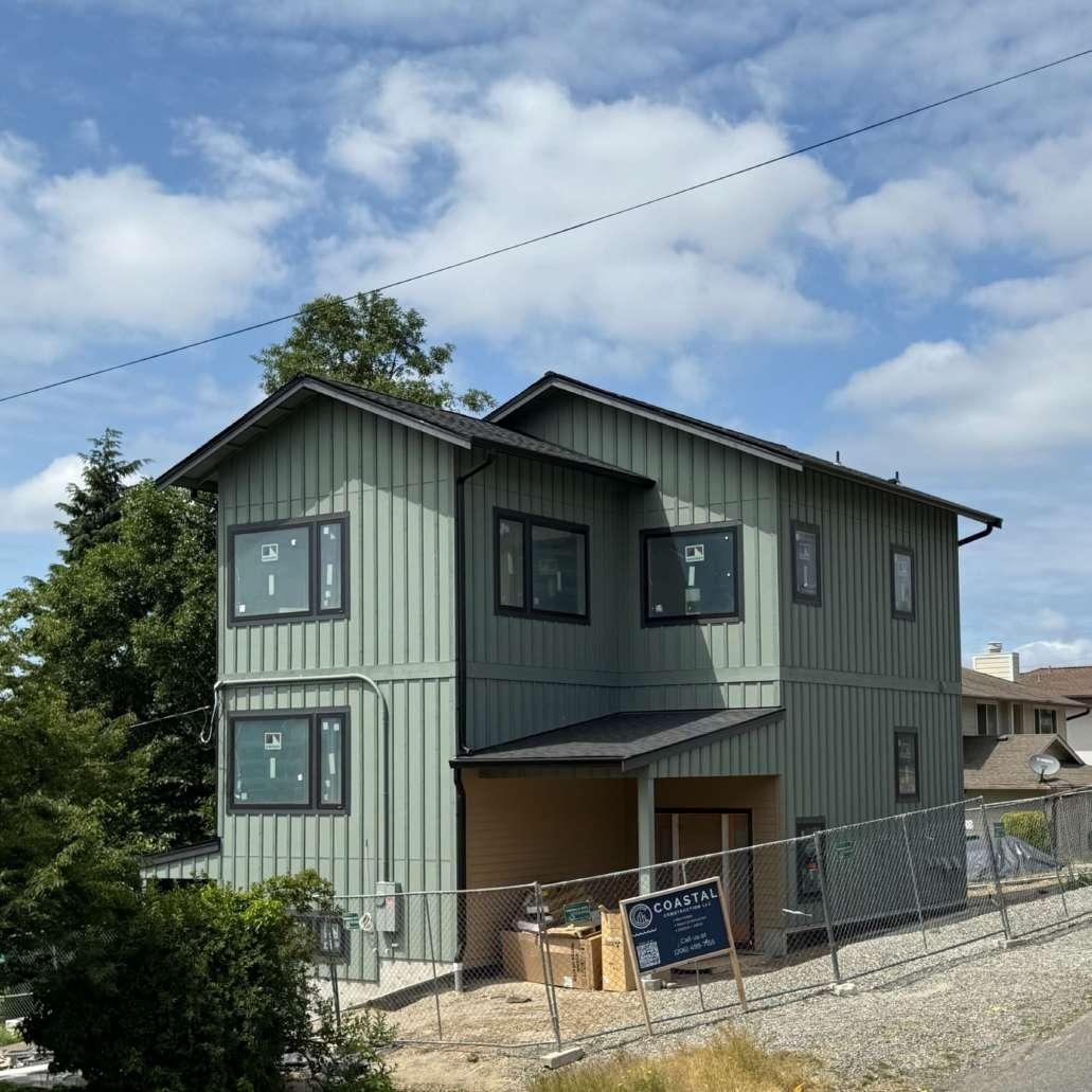 Exterior of a three-story house under construction, with green vertical siding installed, black-trimmed windows, and a construction fence in front