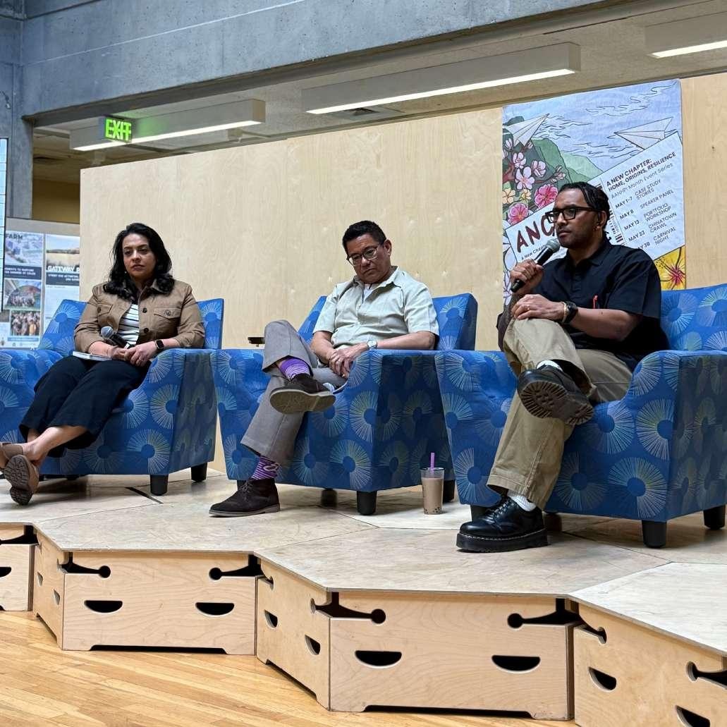 Three panelists seated on blue armchairs on a small stage, one speaking into a microphone, with a poster and 'EXIT' sign visible behind them