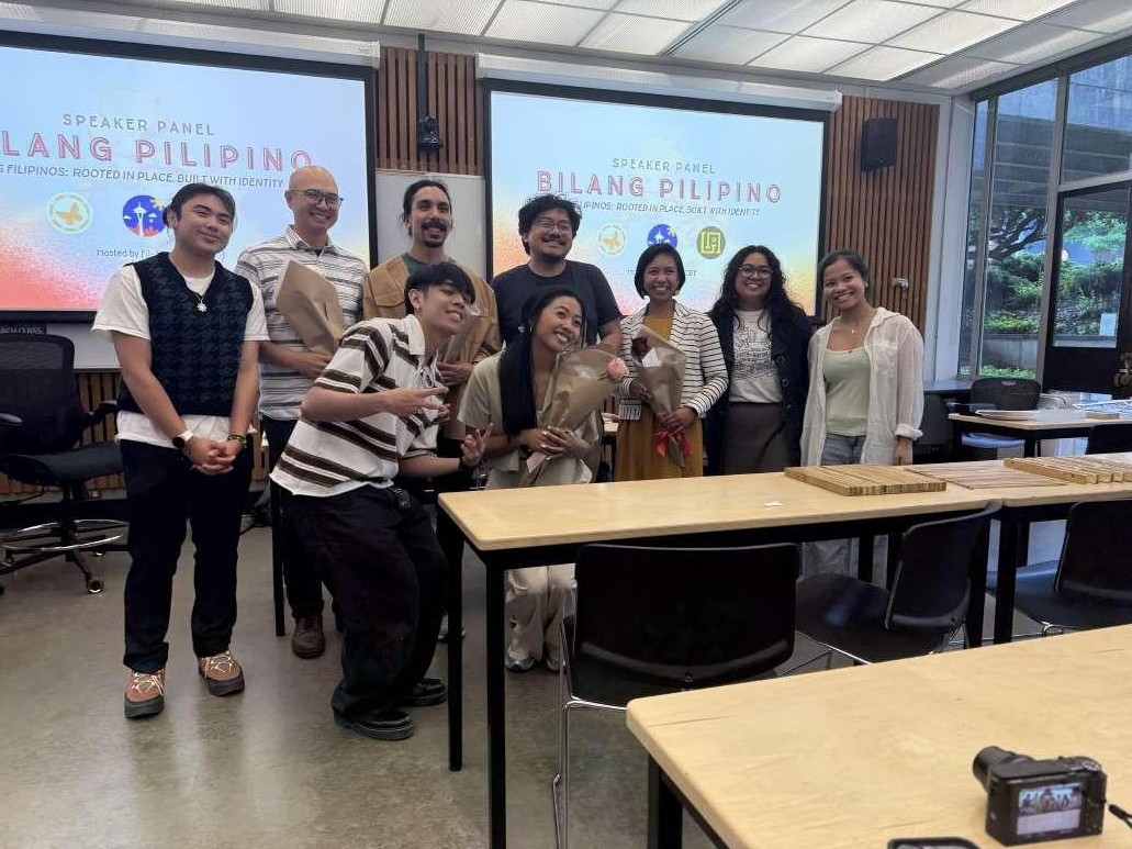 Group of nine people posing and smiling together in a classroom, with two 'Bilang Pilipino' speaker panel screens displayed behind them